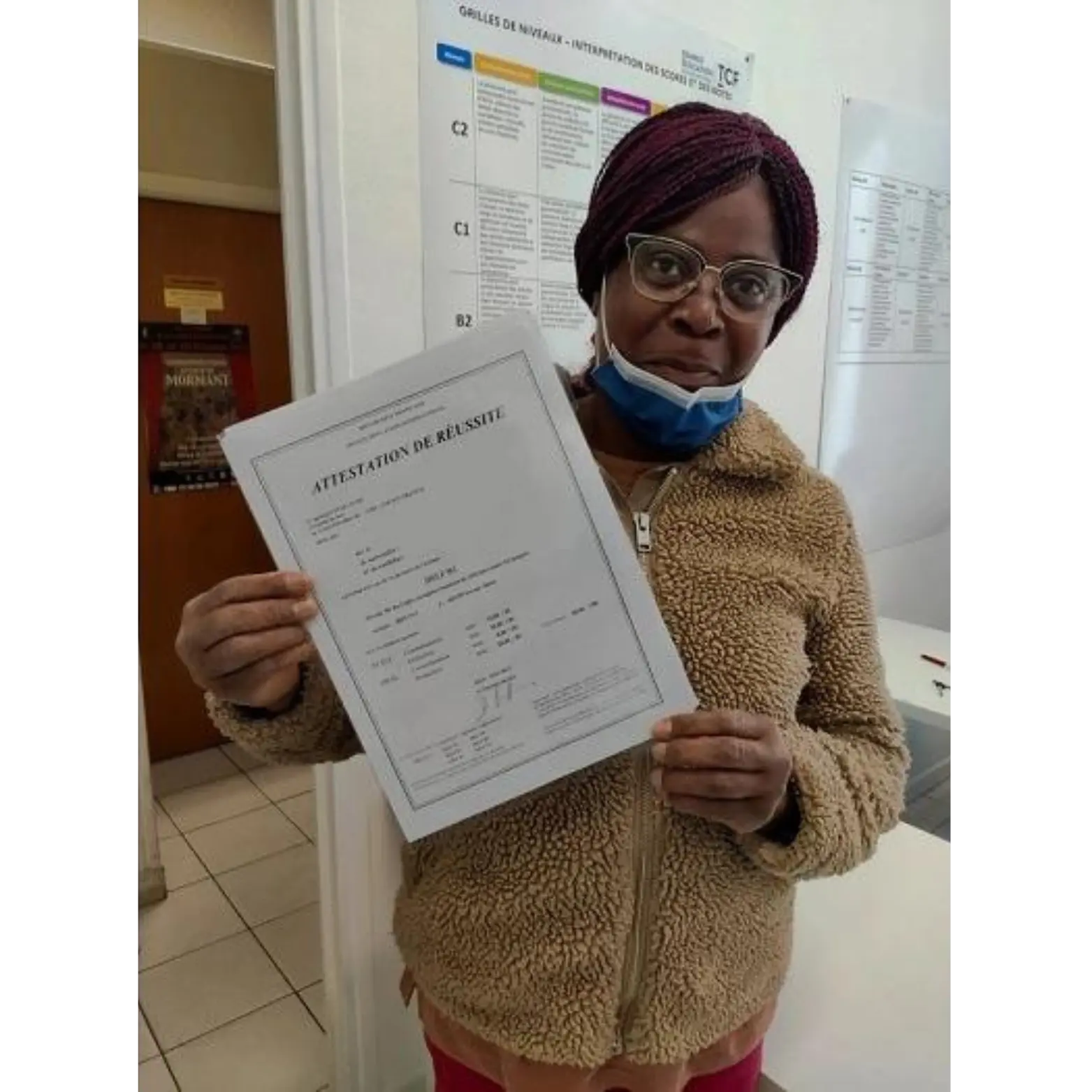 Une femme aux cheveux auburn, portant des lunettes et un manteaux beigne tient fièrement son document d'attestation d'obtention de son diplome de langue