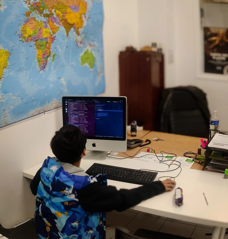 Un enfant, de dos et avec un pull à capuche souris d'ordinateur dans la mains observe un écran d'ordinateur sur lequel est inscrit du code informatique